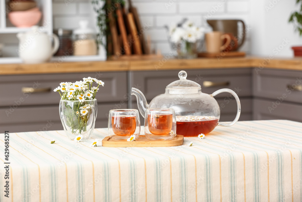 Wooden tray with teapot, cups of natural chamomile tea and flowers on table in kitchen