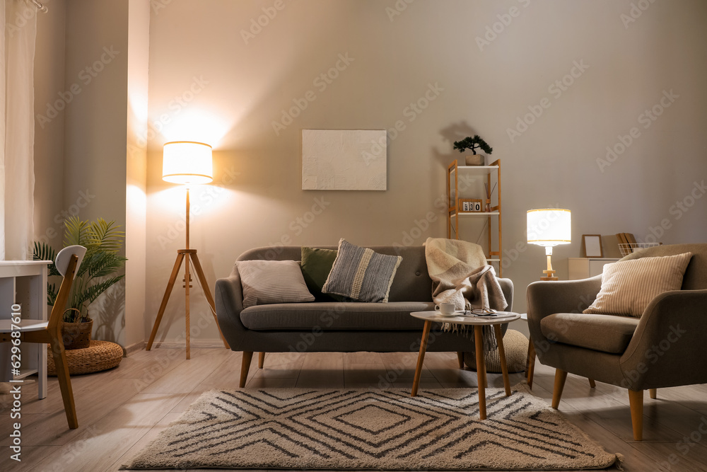 Interior of living room with cozy grey sofa, armchair and glowing lamps