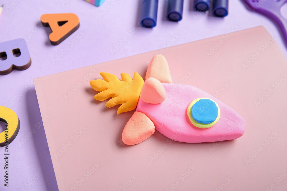 Rocket made of plasticine with different stationery and wooden letters on lilac background