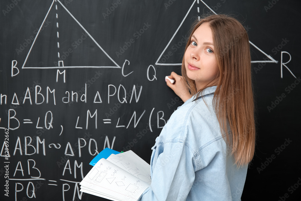 Young math teacher with copybooks writing on blackboard in classroom