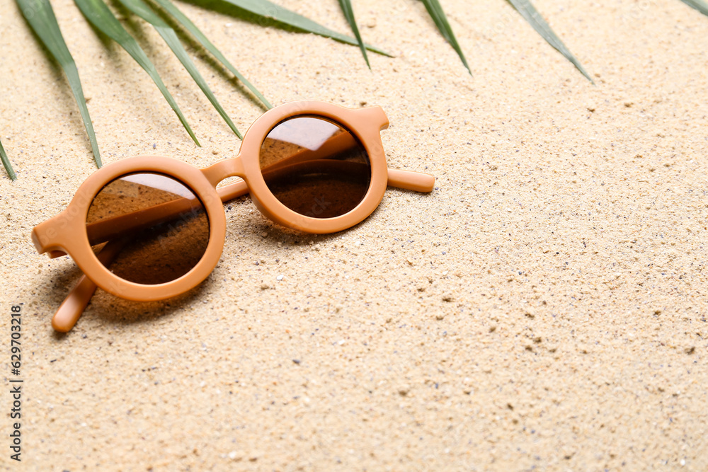 Stylish female sunglasses and palm leaf on sand background