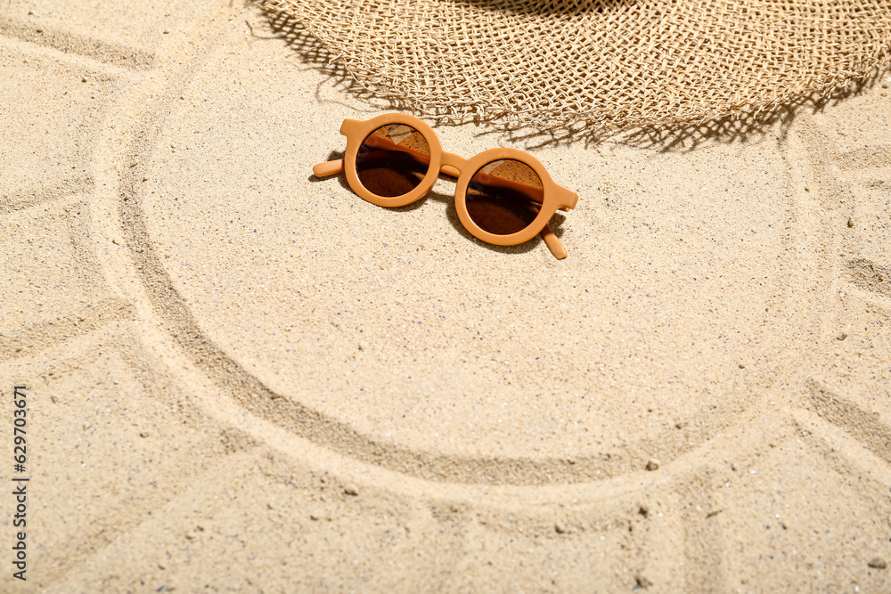 Stylish sunglasses, hat and sun drawn on beach sand