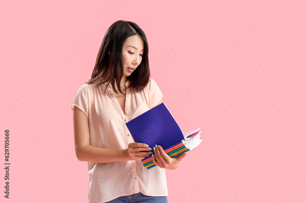 Female Asian teacher with copybooks on pink background