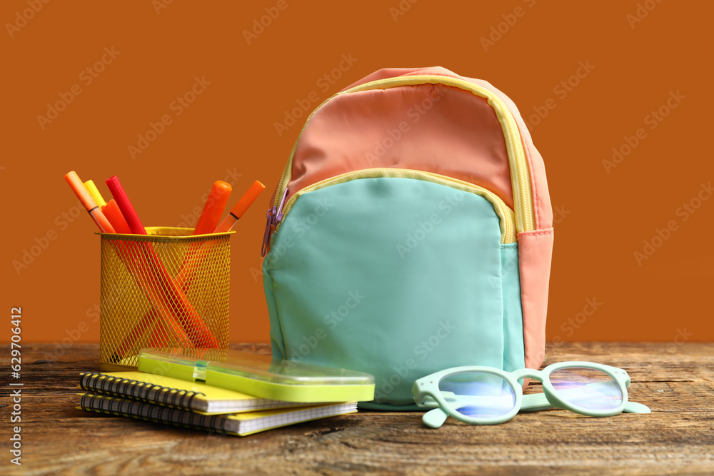 Colorful school backpack with eyeglasses, notebooks and cup of markers on wooden table near orange w