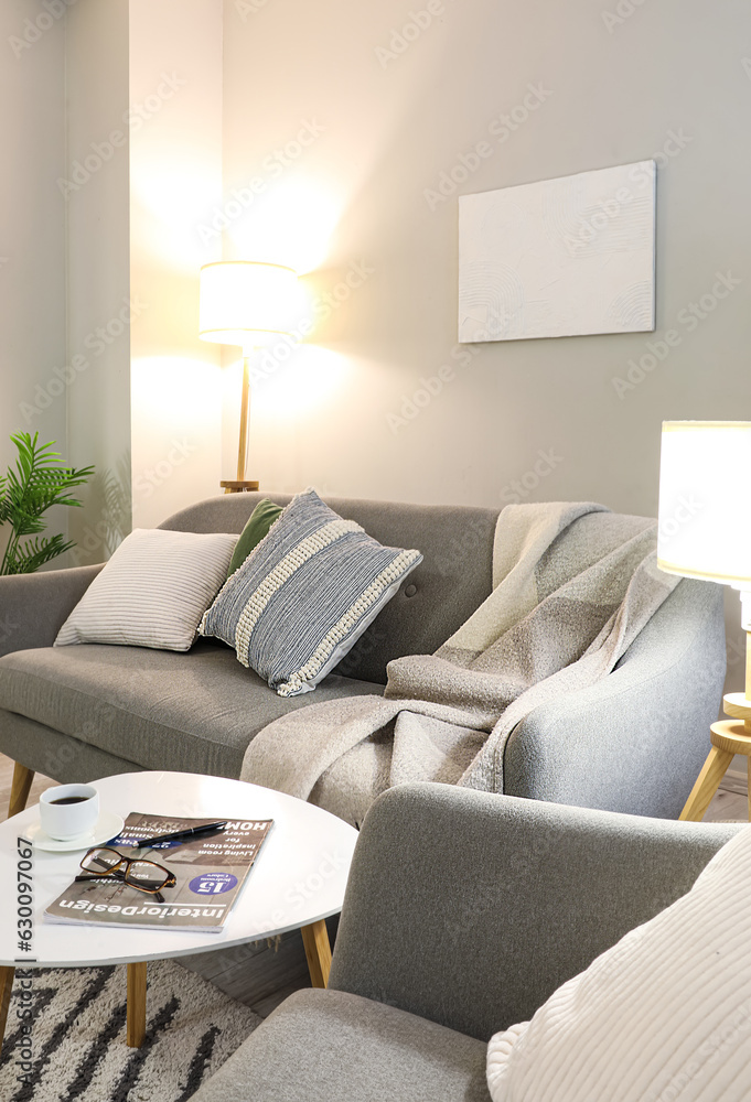 Interior of living room with cozy grey sofa, armchair and glowing lamp