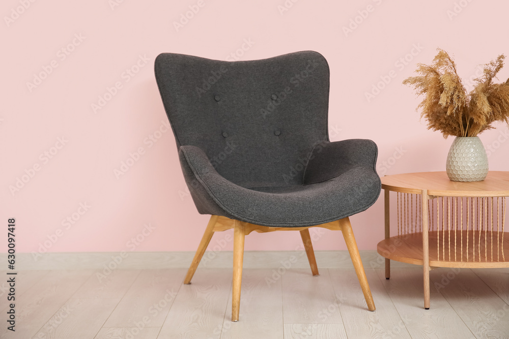 Dark grey armchair with wooden coffee table and dry grass in vase near pink wall