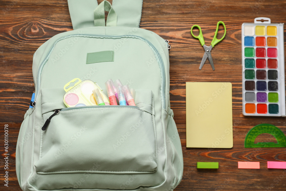 Green school backpack with watercolors, markers and notebooks on wooden background