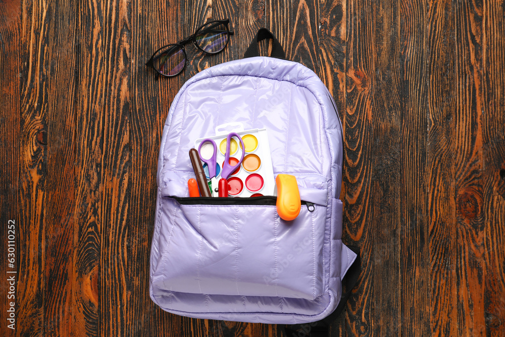 Lilac school backpack with eyeglasses, watercolors and scissors on wooden background