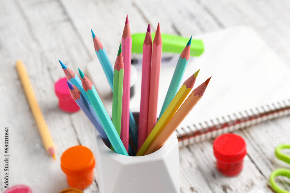 Holder with different color pencils and paints on light wooden background, closeup