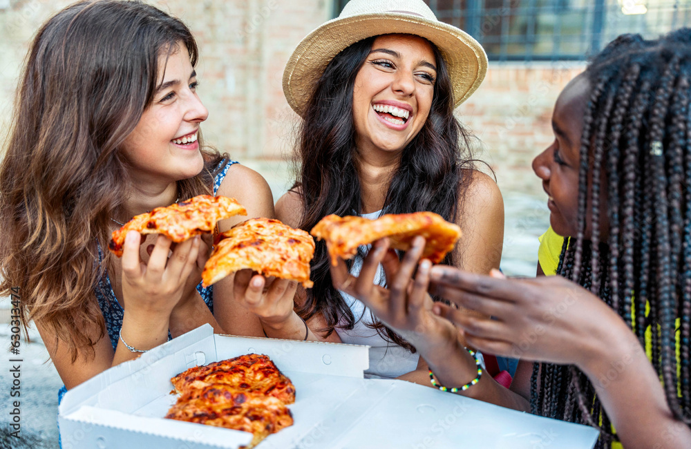 Three young female friends eating pizza sitting outside - Happy women enjoying street food in the ci