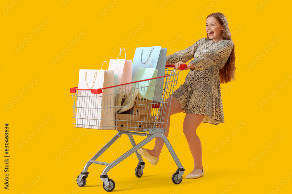 Happy female seller with shopping bags in cart on yellow background