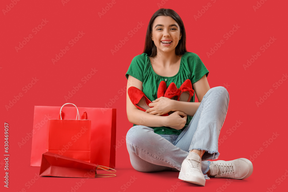 Young woman with stylish shoes and shopping bags sitting on red background