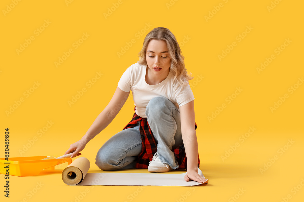 Young woman gluing wallpaper on yellow background