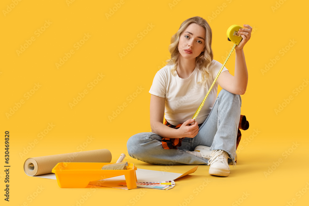 Upset young woman with measuring tape and wallpaper roll on yellow background