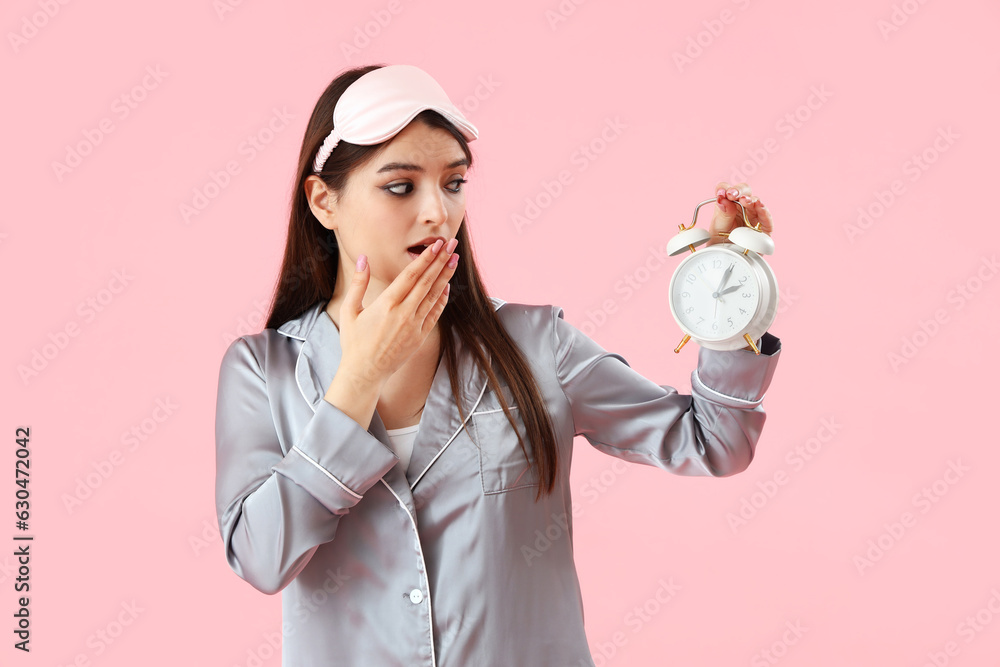 Shocked young woman in pajamas with alarm clock on pink background