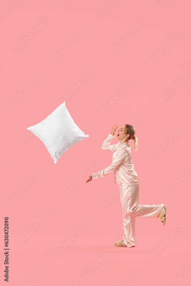 Young woman in pajamas throwing pillow on pink background