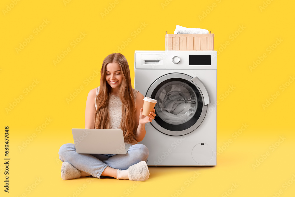 Pretty young woman with modern laptop and cup sitting near washing machine on yellow background