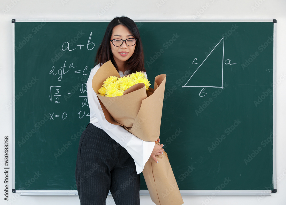 Female Asian teacher with bouquet of flowers near chalkboard in classroom