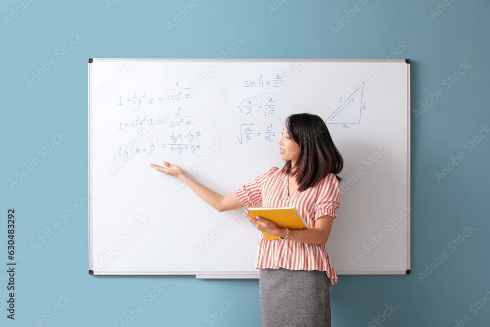 Asian Math teacher with notebook near flipboard in classroom