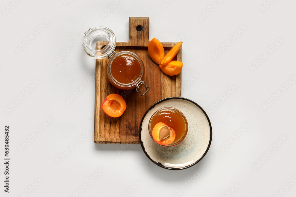 Wooden board with glass bowl and jar of sweet apricot jam on white background