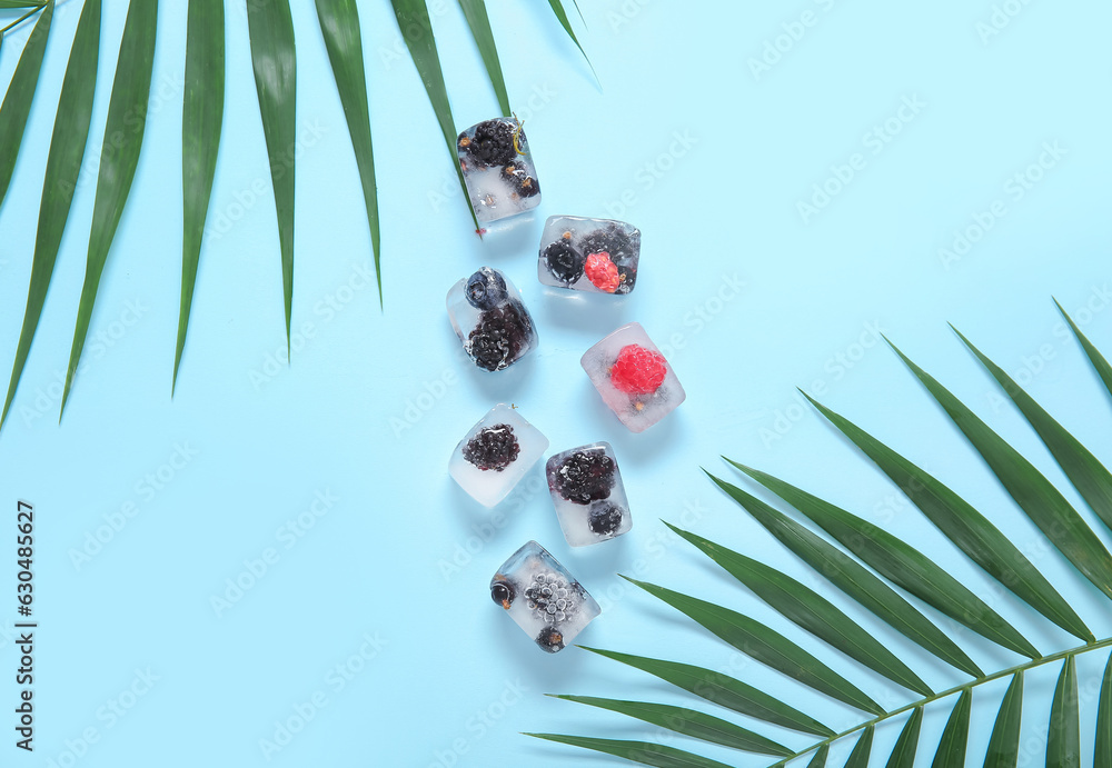 Frozen berries in ice cubes and palm leaves on blue background