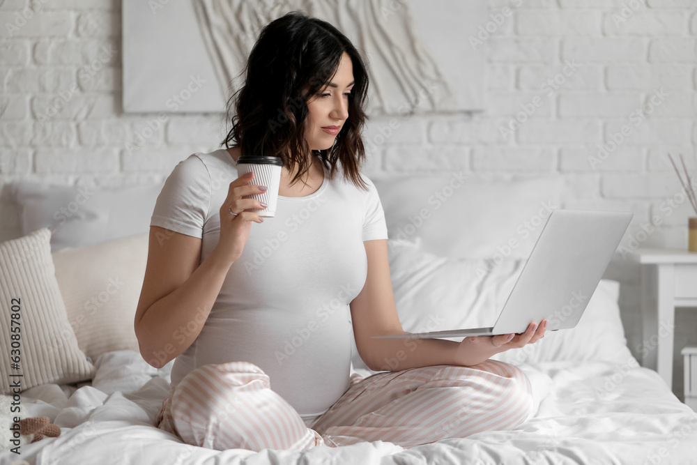 Young pregnant woman with laptop and cup of coffee in bedroom