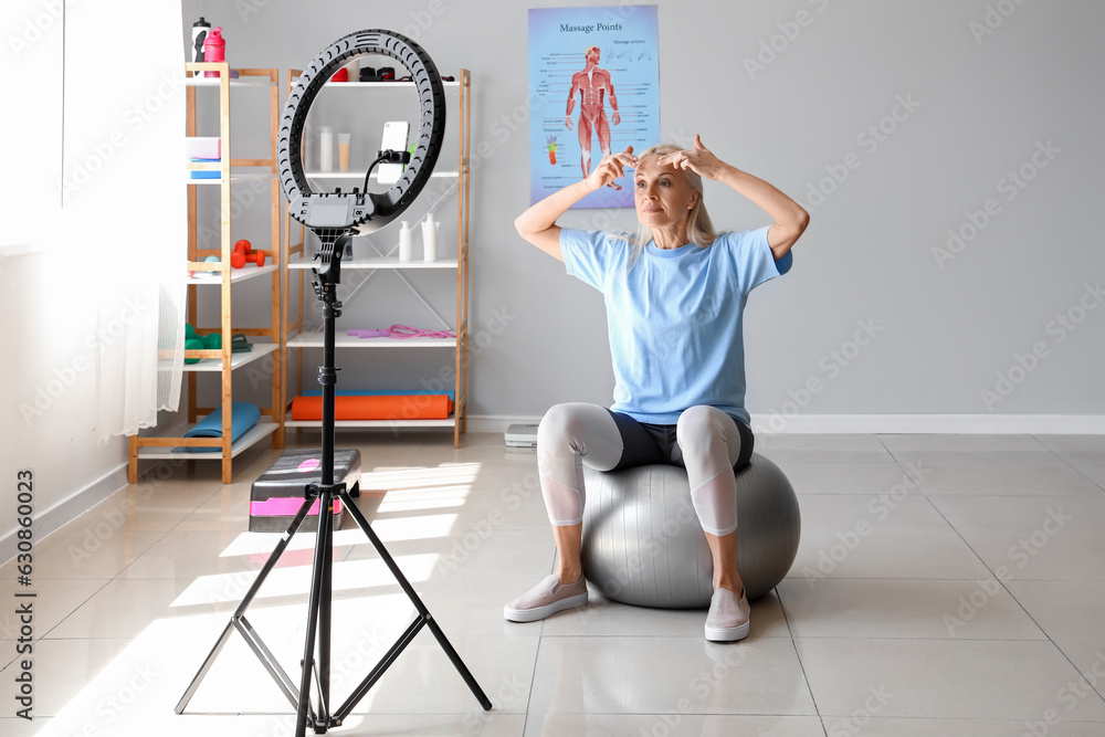 Mature physiotherapist doing face building exercise while recording video in rehabilitation center