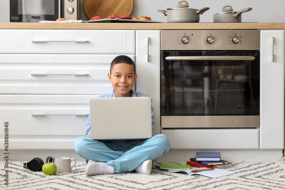 Little African-American boy with laptop studying computer sciences online in kitchen