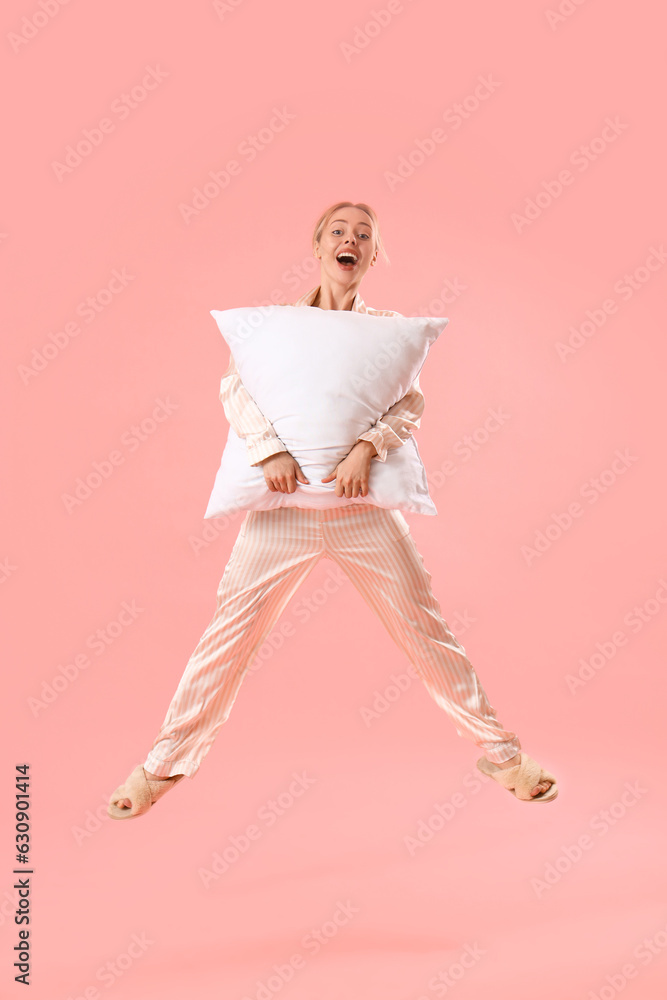 Young woman in pajamas with pillow jumping on pink background