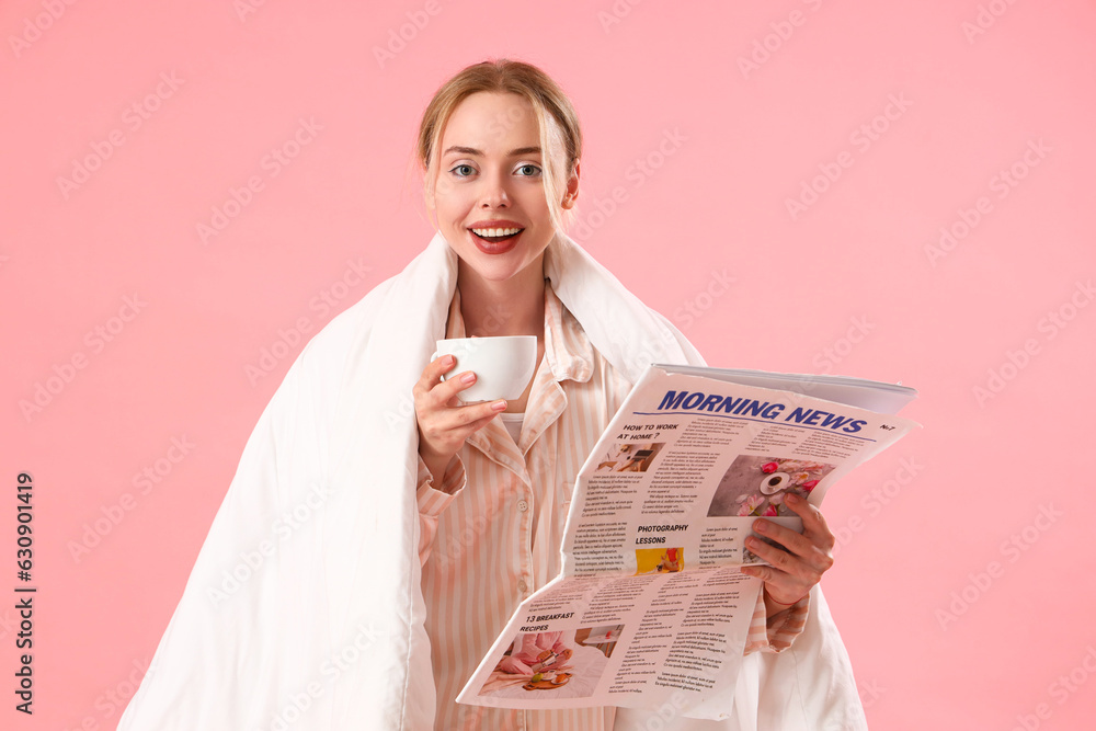 Young woman in pajamas with cup of coffee reading newspaper on pink background