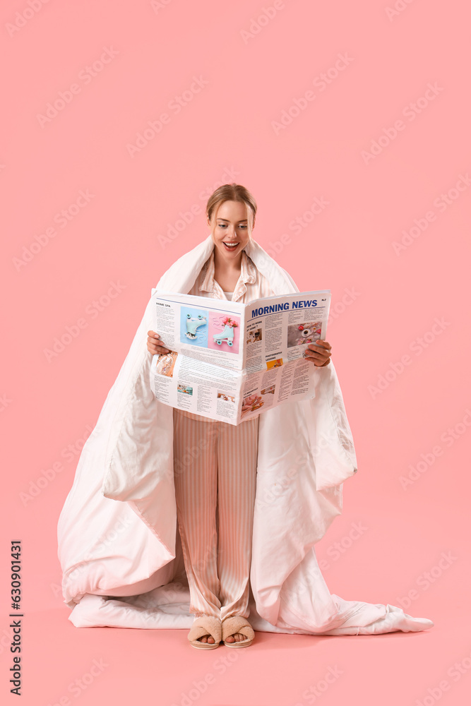 Young woman in pajamas reading newspaper on pink background