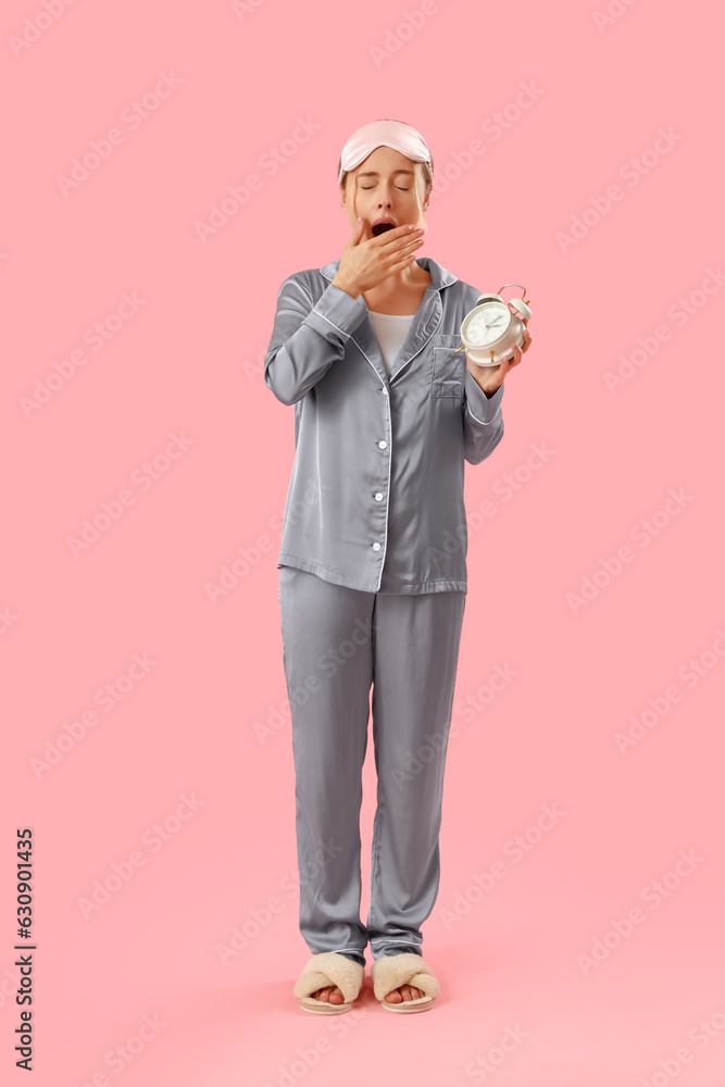 Sleepy young woman in pajamas with alarm clock on pink background