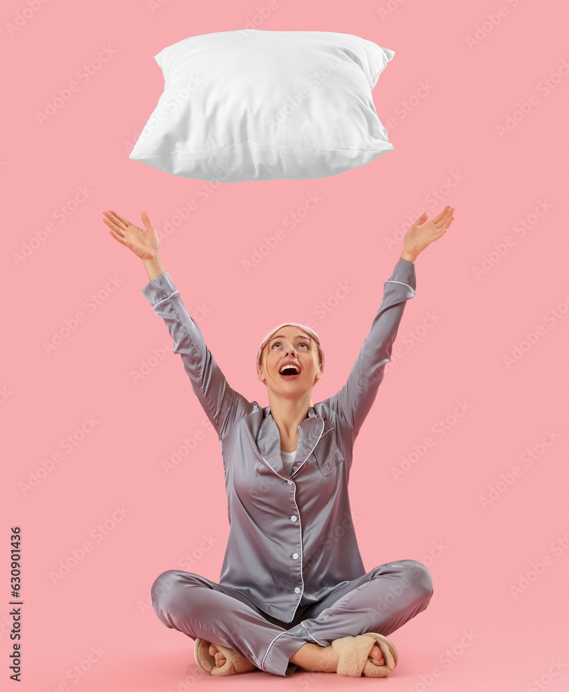 Young woman in pajamas throwing pillow on pink background