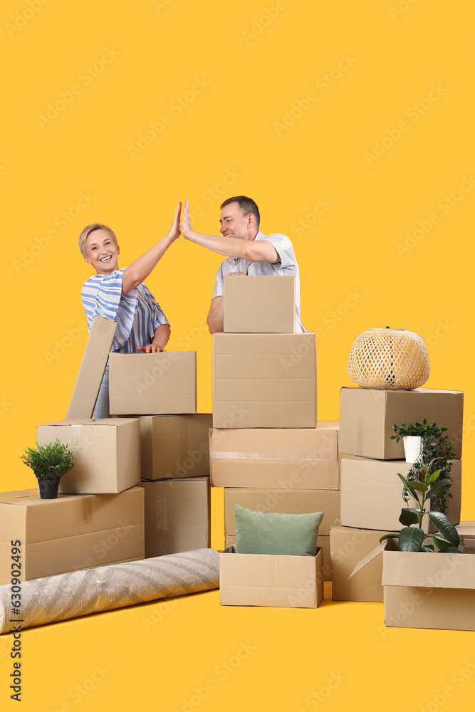 Mature couple with moving boxes giving each other high-five on yellow background