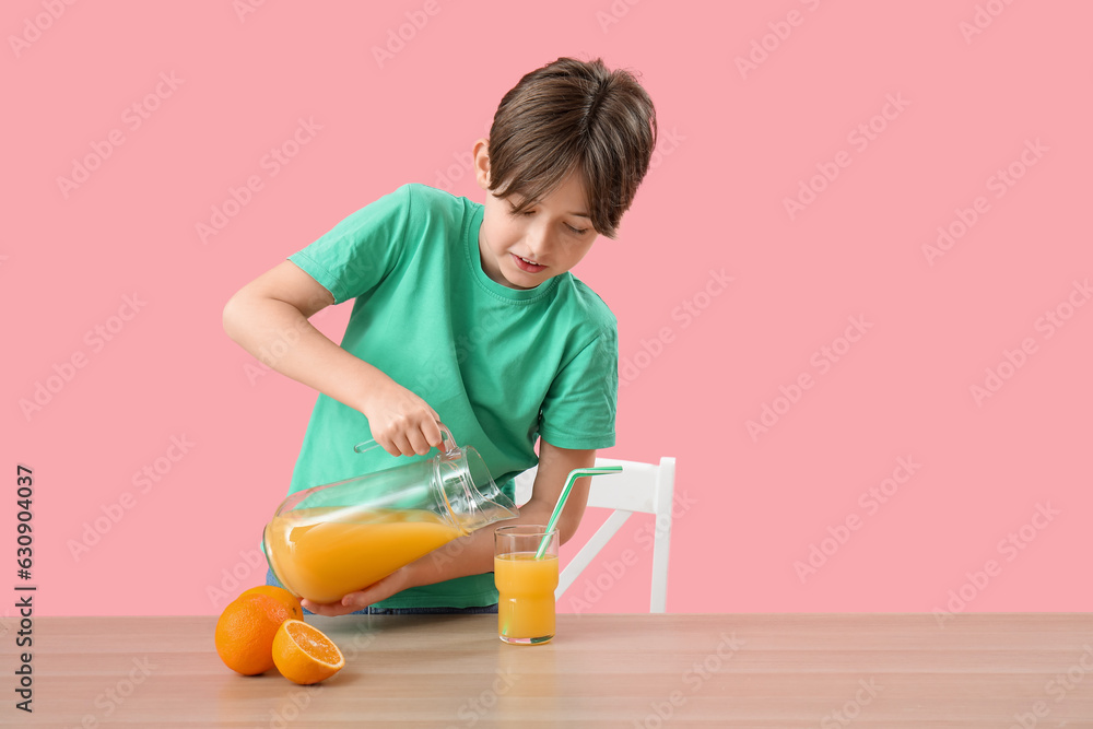 Little boy pouring orange juice in glass at table on pink background