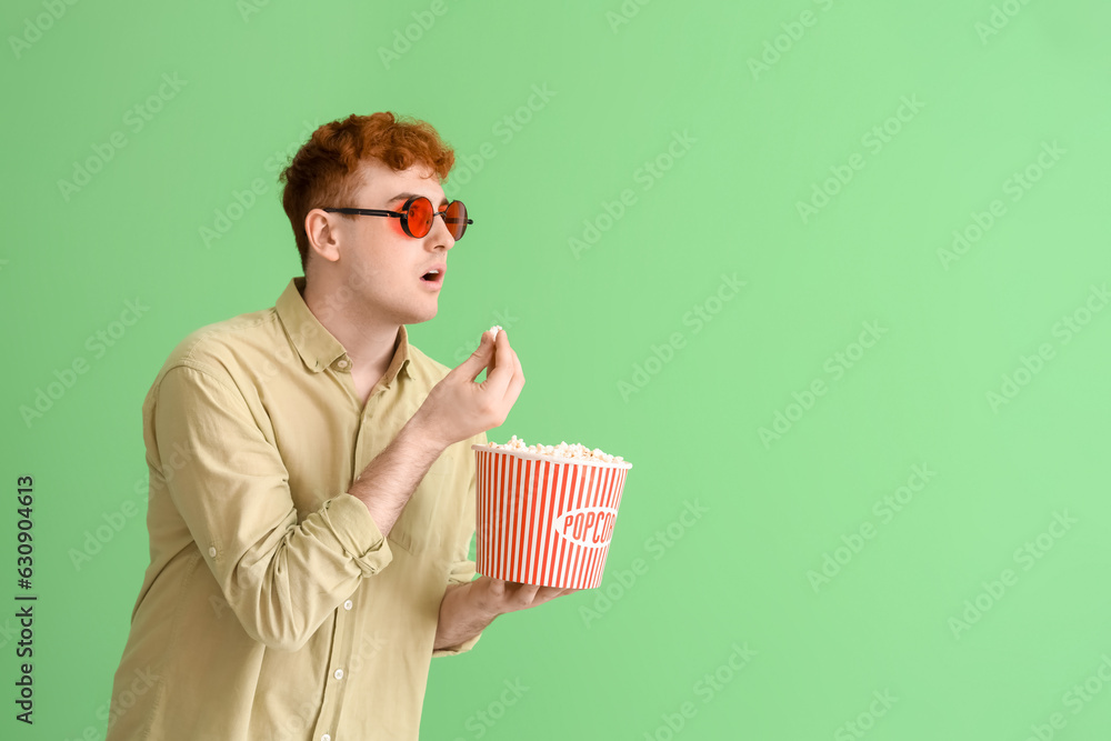 Shocked young redhead man eating popcorn on green background