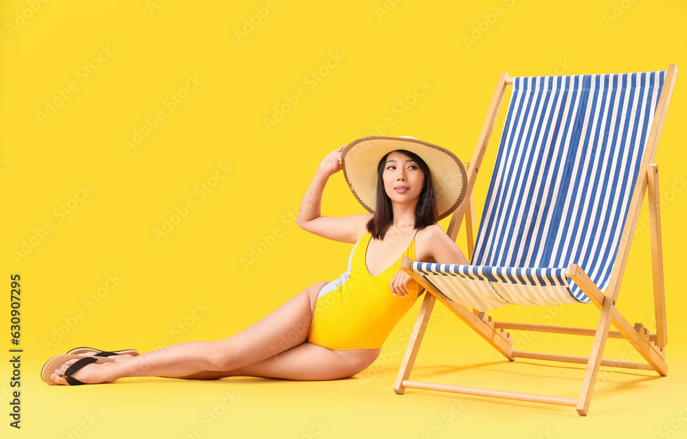 Beautiful Asian woman sitting beside deck chair on yellow background