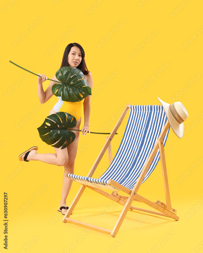 Beautiful Asian woman with palm leaves and deck chair on yellow background