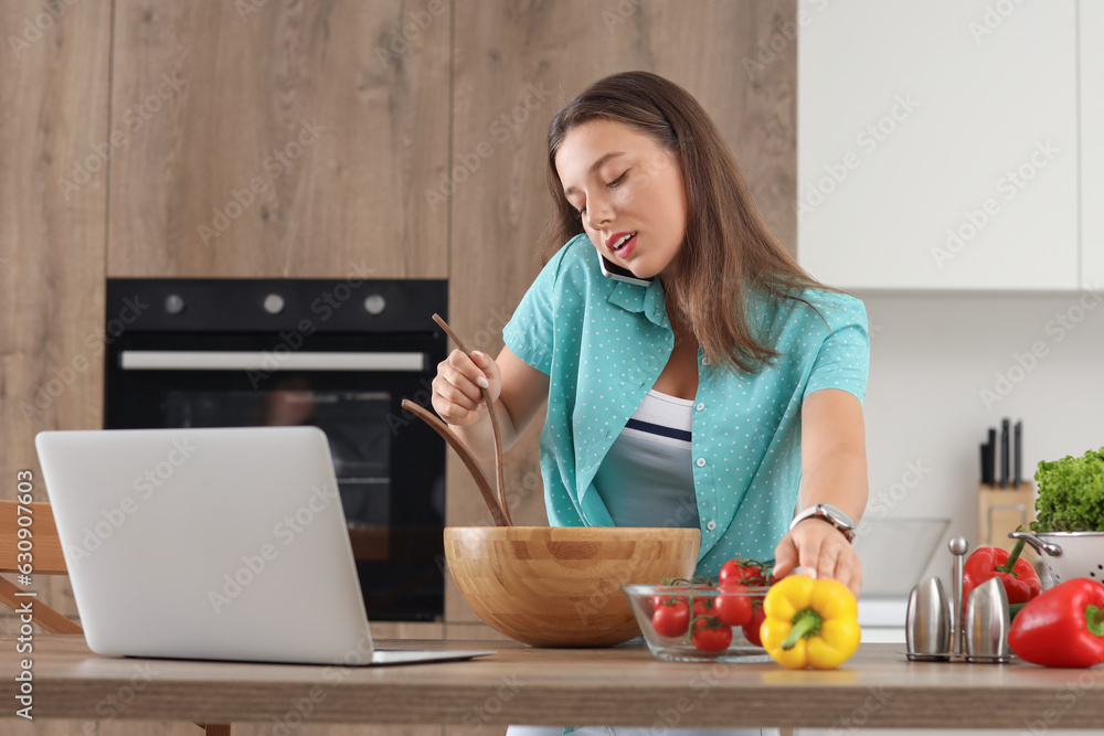 Young woman talking by mobile phone while preparing vegetable salad in kitchen