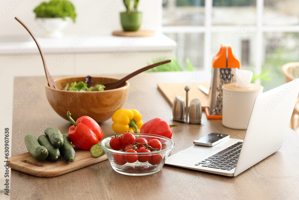 Bowl with vegetable salad and laptop on table in kitchen