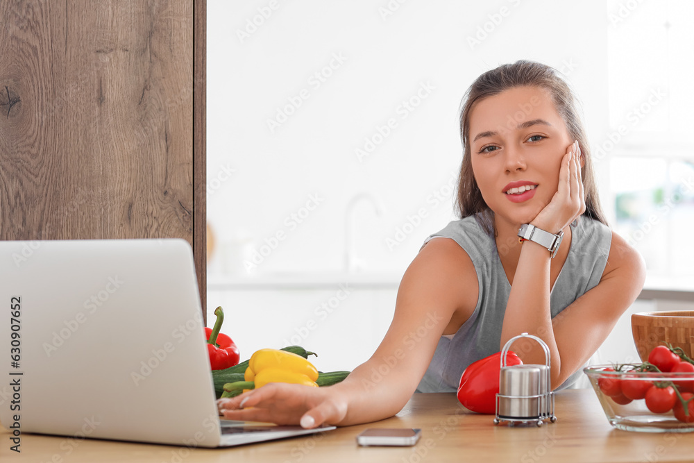 Young woman with laptop and vegetables cooking in kitchen