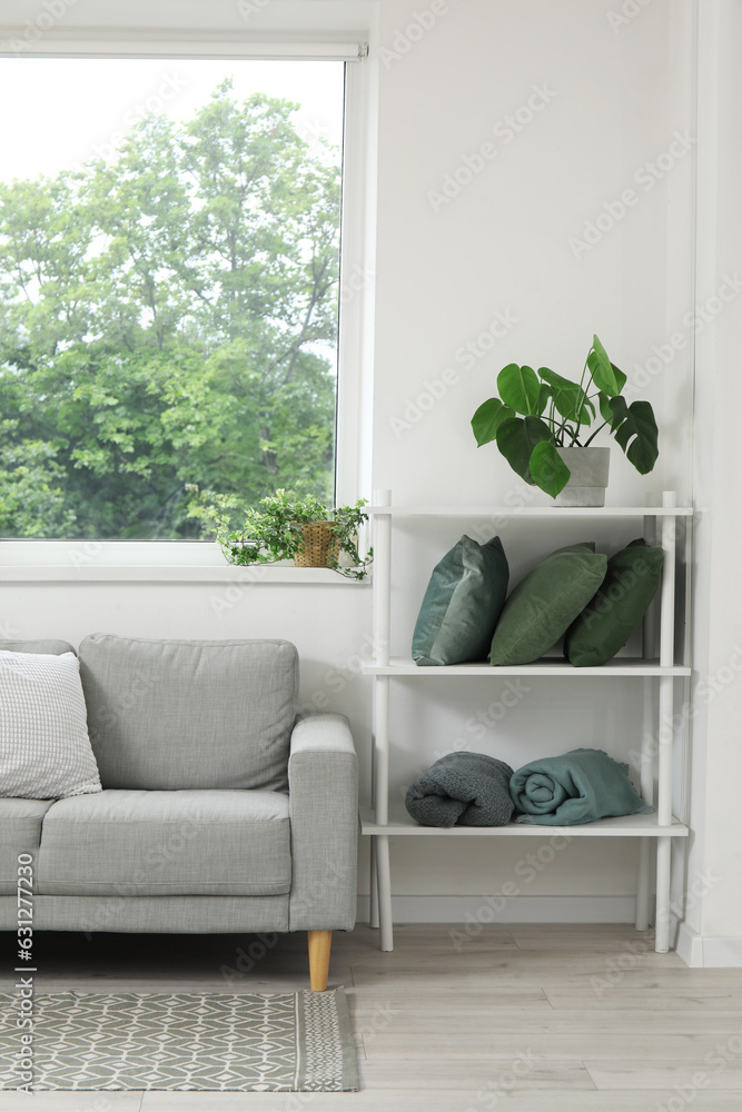 Stylish grey sofa, shelving unit with pillows and houseplants near window in living room