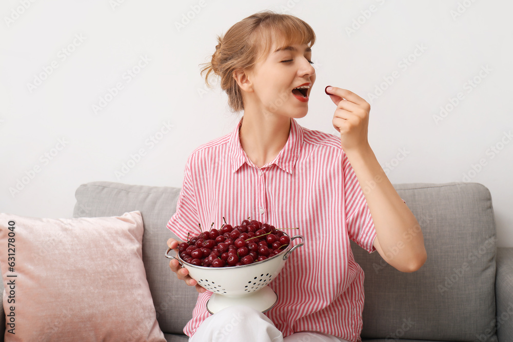 Beautiful happy young woman sitting on sofa and eating ripe cherries in living room