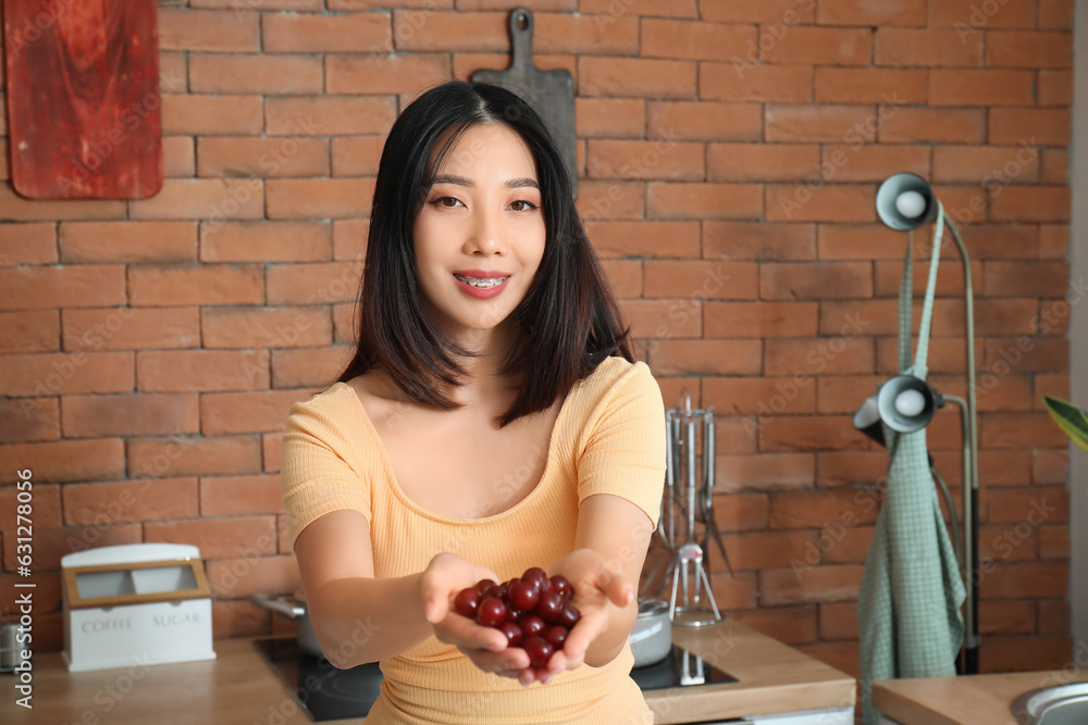 Beautiful young Asian woman holding ripe cherries in kitchen