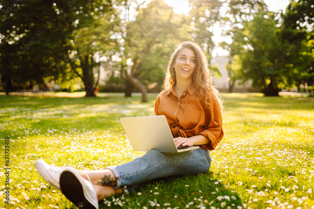 Portrait of a young beautiful female freelancer sitting on a green meadow with a laptop. Education o