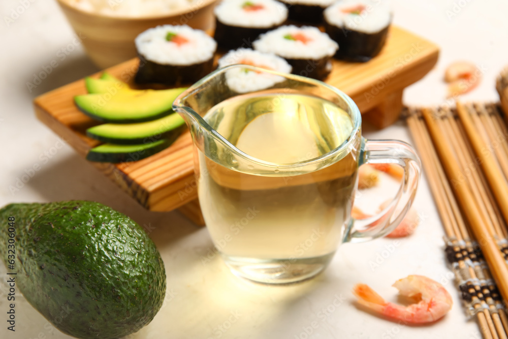 Jug with rice vinegar, avocado and tasty sushi rolls on light background, closeup