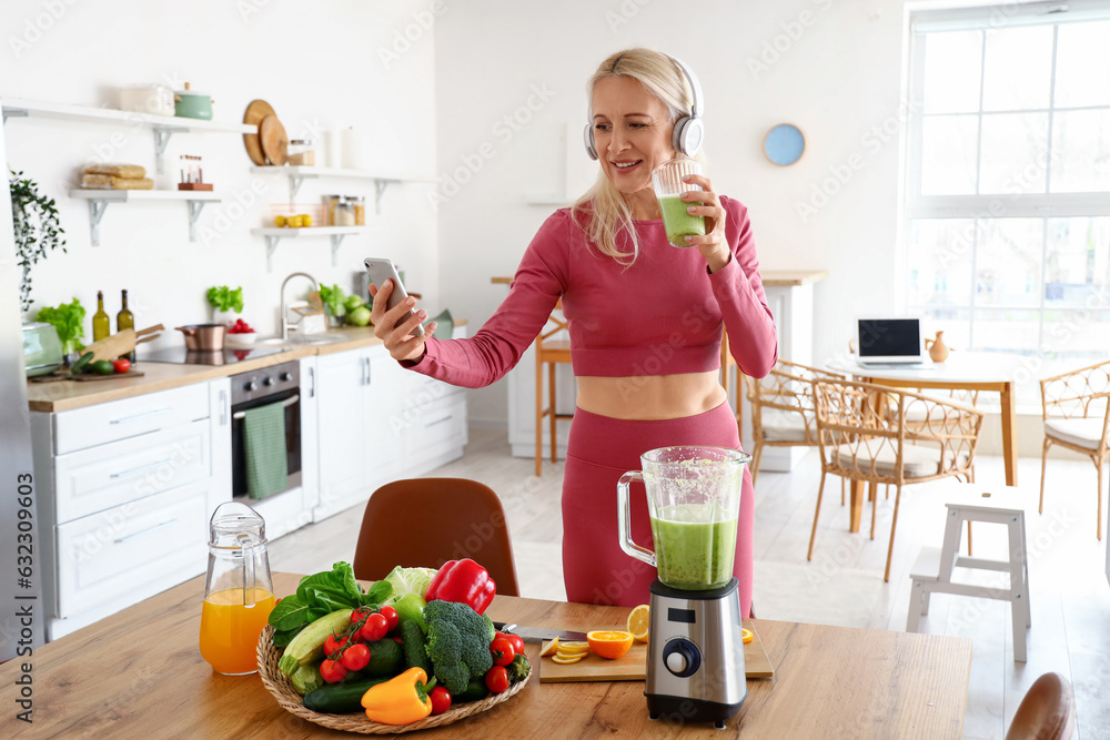 Sporty mature woman with mobile phone and glass of healthy smoothie in kitchen