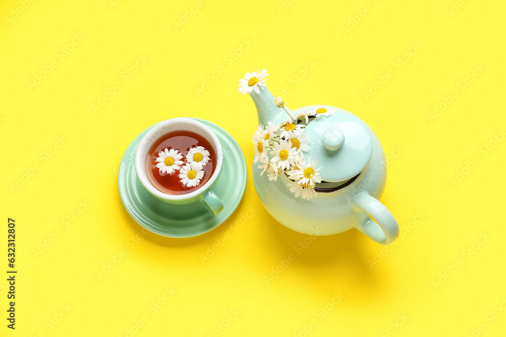 Teapot with cup of natural chamomile tea and flowers on yellow background