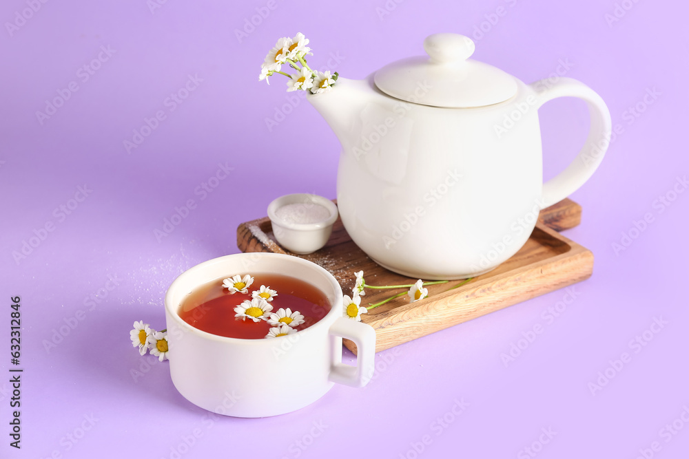 Wooden board with teapot, cup of natural chamomile tea and flowers on lilac background