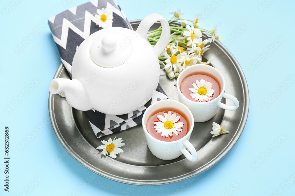 Tray with teapot, cup of natural chamomile tea and flowers on blue background